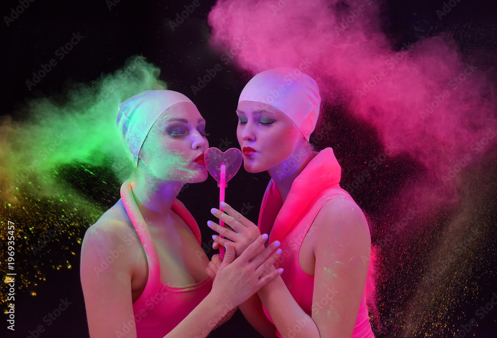 Two beautiful girl feel an affection for each other. They embrace themselves and kiss while multi colored paint powder explodes around them. Two beautiful girl feel an affection for each other. They embrace themselves and kiss while multi colored paint powder explodes around them.