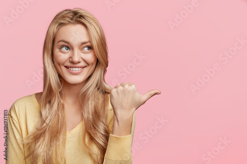 Horizontal portrait of happy female with blonde hair and blue eyes, shing smile, advertizes some product, indicates aside, poses against pink background with copy space for your advertising text