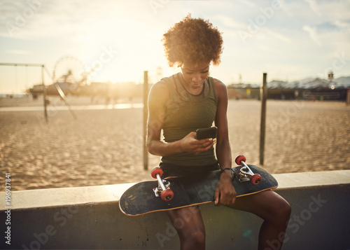 fit african american skater sitting down to look at smart phone at santa monica