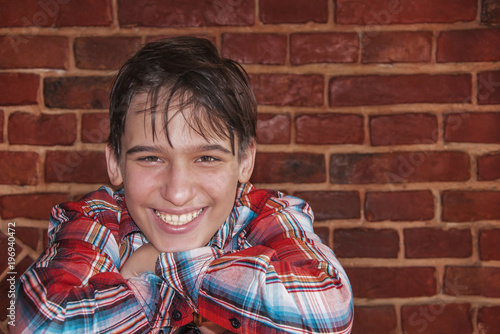 Stylish teenage boy wearing plaid shirt laughing, sitting against red brick wall background (teenager concept human behavior)
