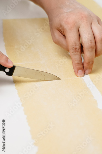 Man's hands cutting thinly rolled layer of Italian pasta dough for making meat or spinach and ricotta filled Ravioli at home