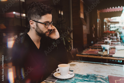 Young handsome serious bearded man entrepreneur in glasses is having work conversation via smartphone while sitting alone in street cafe with the cup of a hot delicious coffee on the table
