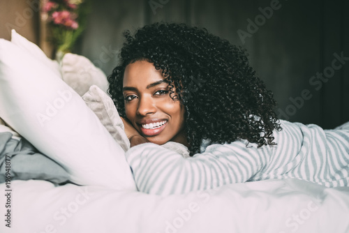 close up of a pretty black woman with curly hair smiling and lying on bed looking at the camera
