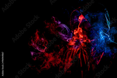 A young man in a black hood smokes and breathes out the colored blue and red  smoke from the vape on a black  isolated background