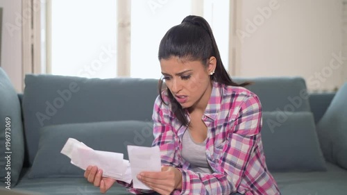 lateral panning of young attractive worried hispanic woman checking bills bank papers expenses and monthly payments stressed sititing at apartment couch in domestic stress finance problem 