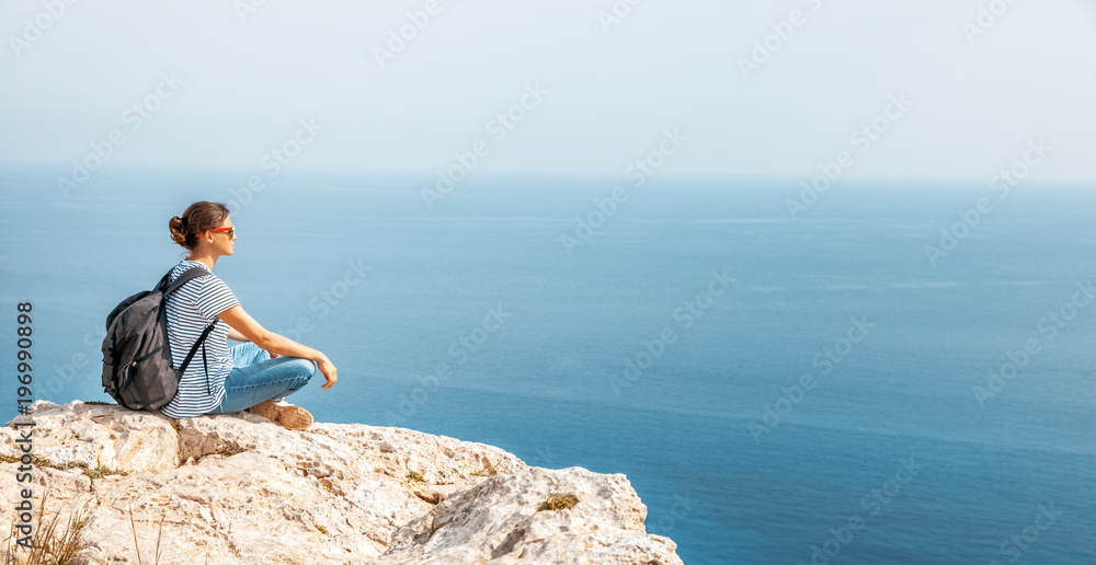 A girl traveler sits on a rock and admires the blue boundless sea, freedom, travel, unity with nature,