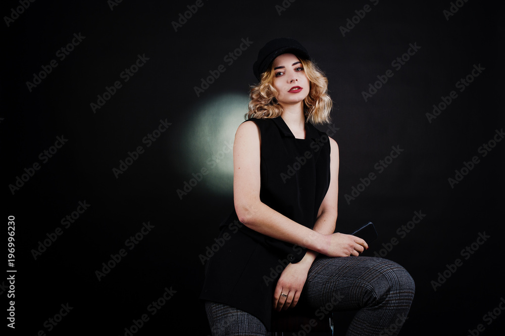 Studio portrait of blonde girl in black wear and cap with mobile phone at hand against dark background. Studio portrait of blonde girl in black wear and cap with mobile phone at hand against dark background.