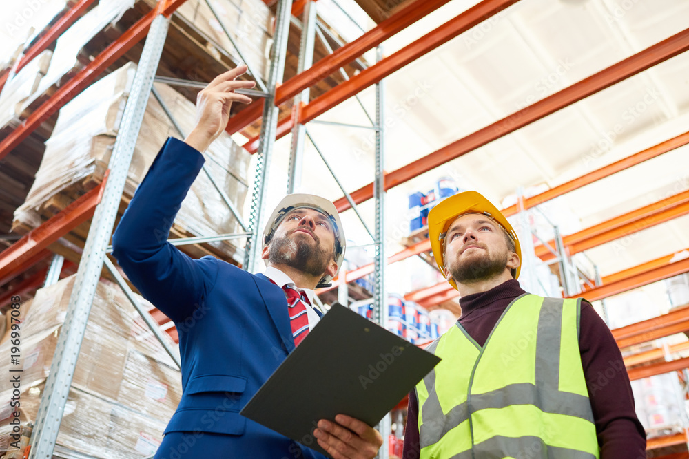 Low angle portrait of warehouse manager holding clipboard talking to worker wearing hardhat and reflective jacket while discussing stock inventory