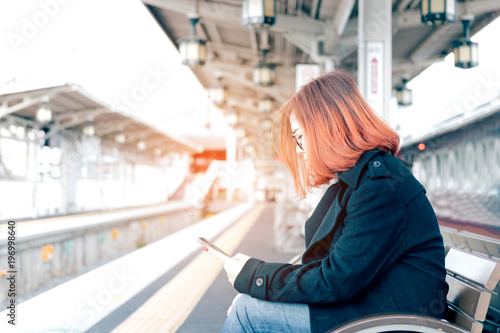 Young asian woman sitting on bench waiting for train and using mobile phone.