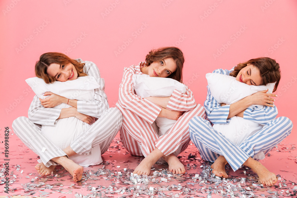 Photo of sleepy women 20s wearing leisure clothings holding pillows and taking pleasure during slumber party, isolated over pink background - obrazy, fototapety, plakaty 