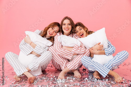 Photo of happy women 20s wearing leisure clothings sitting barefoot on floor with legs crossed and hugging pillows during girlish sleepover, isolated over pink background