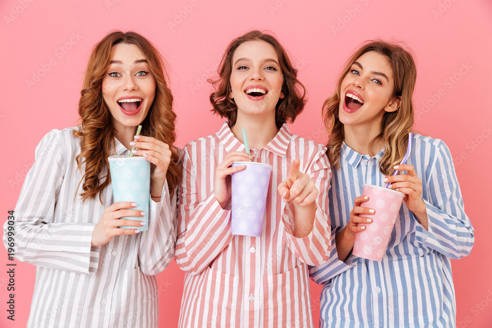 Obraz premium Three beautiful young girls 20s wearing colorful striped pyjamas smiling and drinking cold soda from paper cups during happy sleepover, isolated over pink background