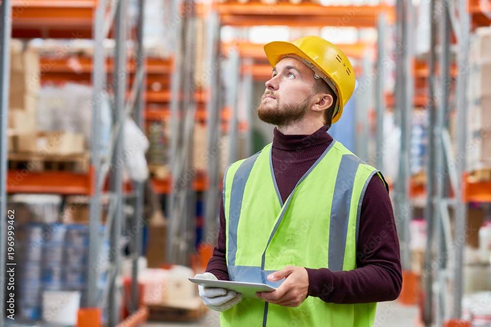 Waist up portrait of modern warehouse worker using digital tablet looking up at tall shelves, copy space Waist up portrait of modern warehouse worker using digital tablet looking up at tall shelves, copy space