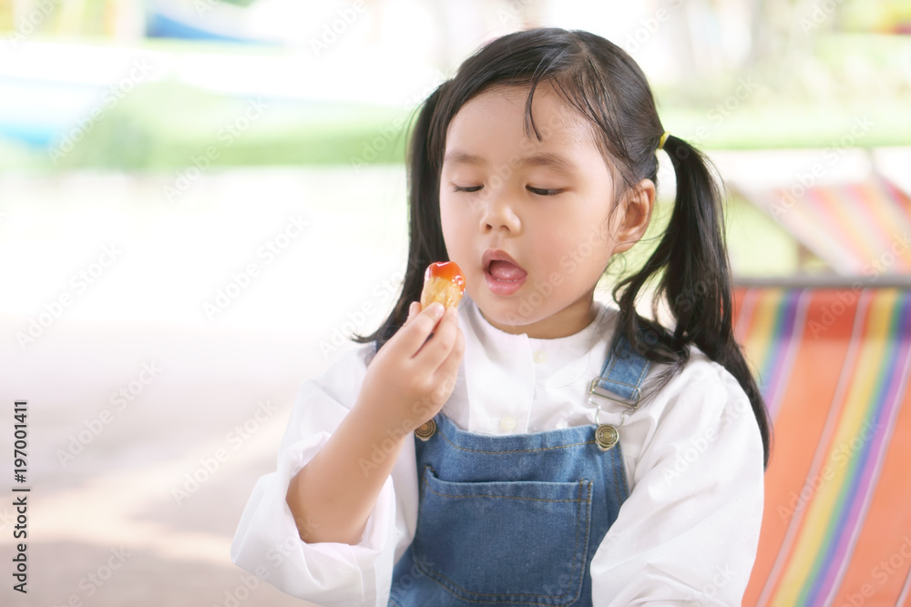 Asian children cute or kid girl enjoy eating sausages with tomato sauce or ketchup and wear overalls or jumper for lunch food or dessert because hungry and hot weather with sweat on summer holiday Asian children cute or kid girl enjoy eating sausages with tomato sauce or ketchup and wear overalls or jumper for lunch food or dessert because hungry and hot weather with sweat on summer holiday