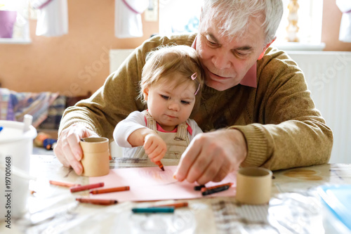 Cute little baby toddler girl and handsome senior grandfather painting with colorful pencils at home. Grandchild and man having fun together photo