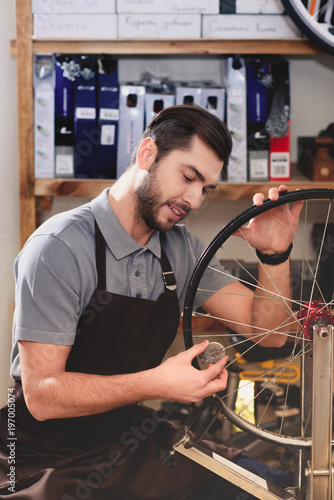 smiling young man in apron fixing bicycle wheel in workshop