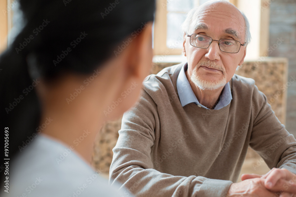 Social interaction. Handsome positive senior man wearing glasses while listening to volunteer and posing on blurred background Social interaction. Handsome positive senior man wearing glasses while listening to volunteer and posing on blurred background