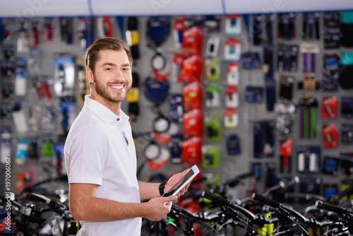 handsome young worker holding digital tablet and smiling at camera in bike shop