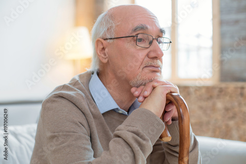 Depressive mood. Handsome reflective senior man leaning on cane while wearing glasses and posing on blurred background