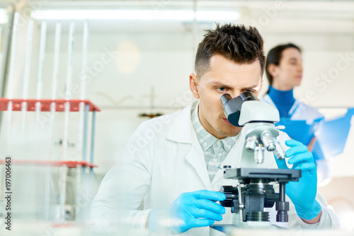 Handsome young scientist with stylish haircut using microscope while working on ambitious project  interior of modern laboratory on background