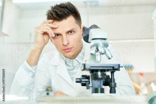 Portrait shot of handsome young researcher with stylish haircut sitting at laboratory bench and looking at camera, modern microscope on foreground