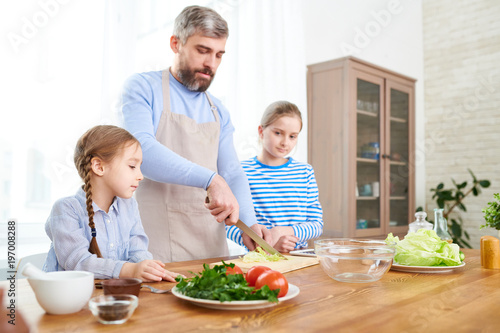 Portrait of mature bearded man teaching two girls cooking in kitchen at home  cutting vegetables and making salad