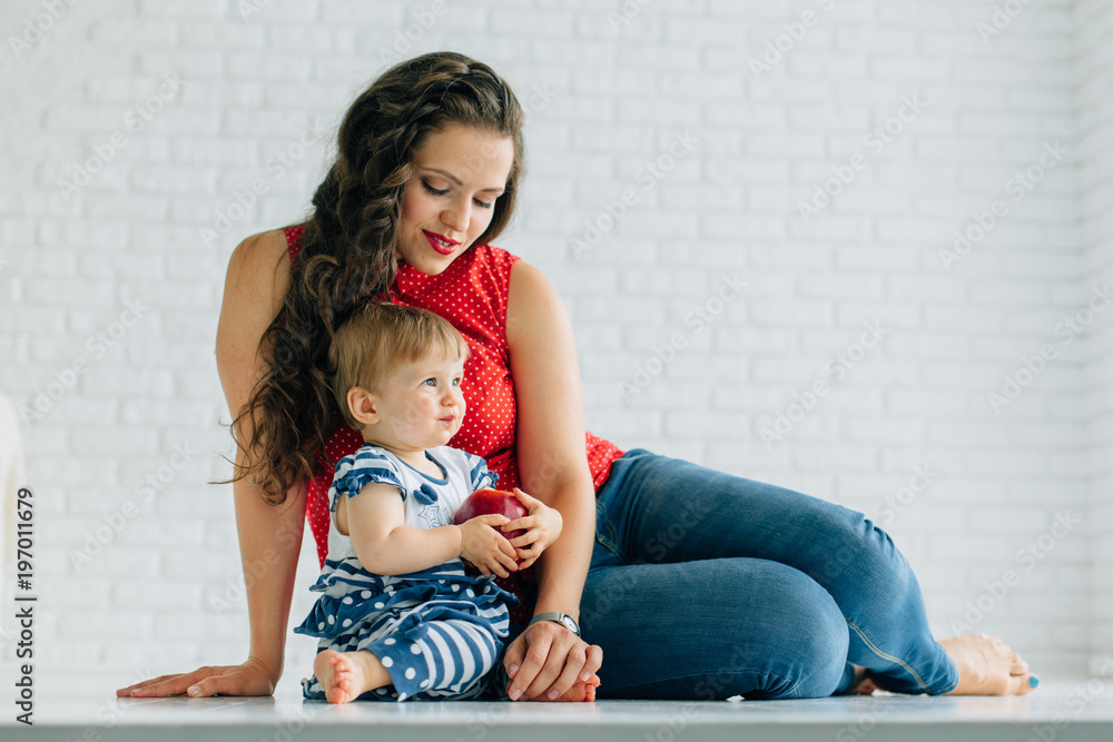 Happy mother with little baby girl. Woman with long dark curly hair in jeans and red blouse with white polka dots and daughter with red apple. White living room Happy mother with little baby girl. Woman with long dark curly hair in jeans and red blouse with white polka dots and daughter with red apple. White living room