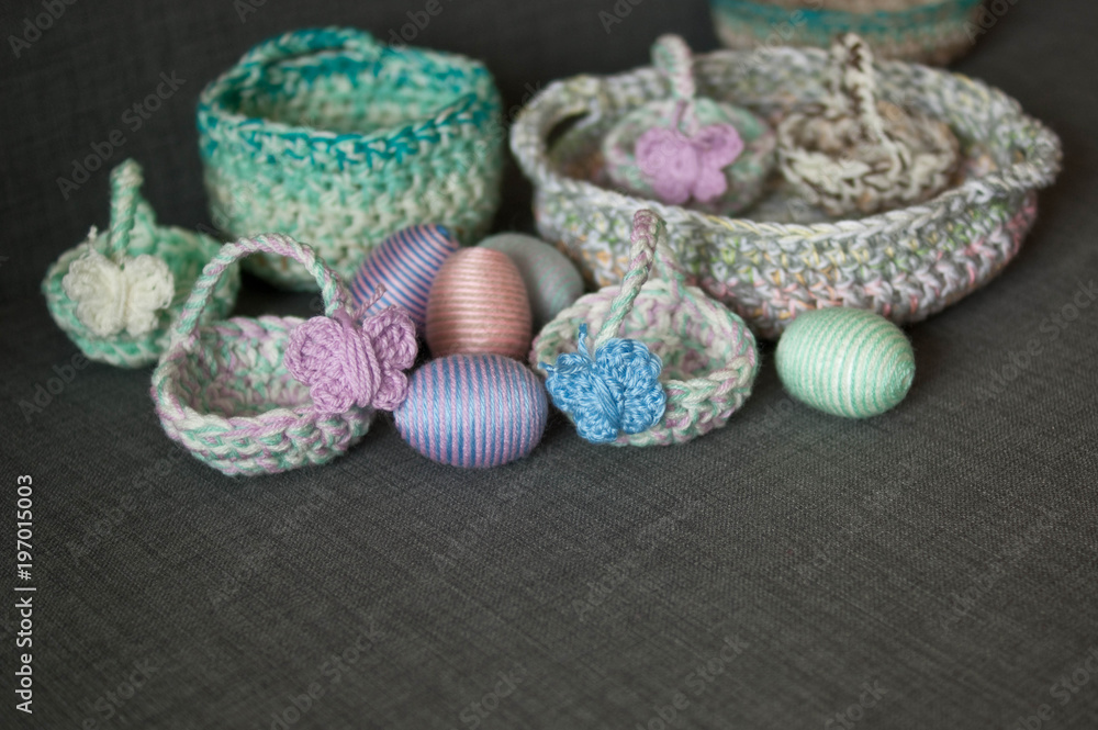 Fototapeta premium Easter decoration, crochet basket, striped colorful egg wrapped in soft yarn thread, pastel colors, butterfly, dark grey texture background. Homemade decor. Shallow depth of focus. Easter holidays.