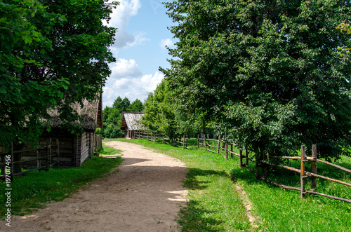 Old village street in sunny summer day with ground pathway, green grass and trees, and traditional pole fence