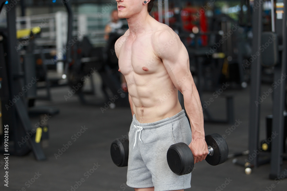 A shirtless young muscular man working out in gym doing exercises with equipment in fitness center. A shirtless young muscular man working out in gym doing exercises with equipment in fitness center.