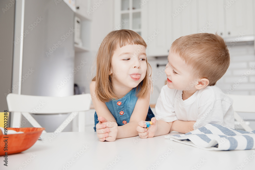 Children boy and girl are sitting at white dining table smiling, laughing and looking each other, waiting for breakfast. In foreground eggs. Concept healthy food. Children boy and girl are sitting at white dining table smiling, laughing and looking each other, waiting for breakfast. In foreground eggs. Concept healthy food.
