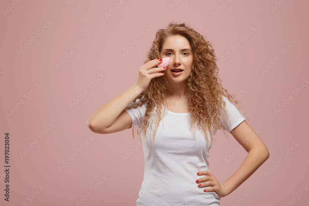 Beautiful girl with beauty blender sponge for make-up, curly hair, on pink background. concept of applying cosmetics to teenagers and adults Beautiful girl with beauty blender sponge for make-up, curly hair, on pink background. concept of applying cosmetics to teenagers and adults