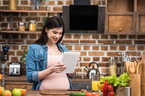 smiling young pregnant woman using digital tablet while cooking at kitchen