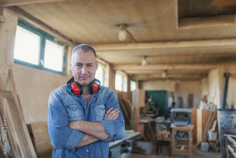 Portrait of a bearded man who owns a small carpentry business, standing in his workshop with with arms folded and showing strong forearms, smiling confidently at the camera Portrait of a bearded man who owns a small carpentry business, standing in his workshop with with arms folded and showing strong forearms, smiling confidently at the camera