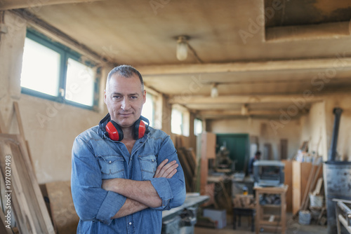 Portrait of a bearded man who owns a small carpentry business, standing in his workshop with with arms folded and showing strong forearms, smiling confidently at the camera © Dragana Gordic