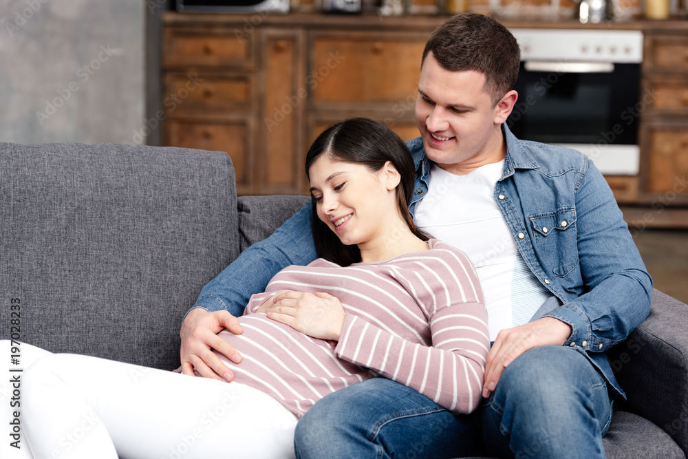 happy young pregnant couple embracing and touching belly together at home