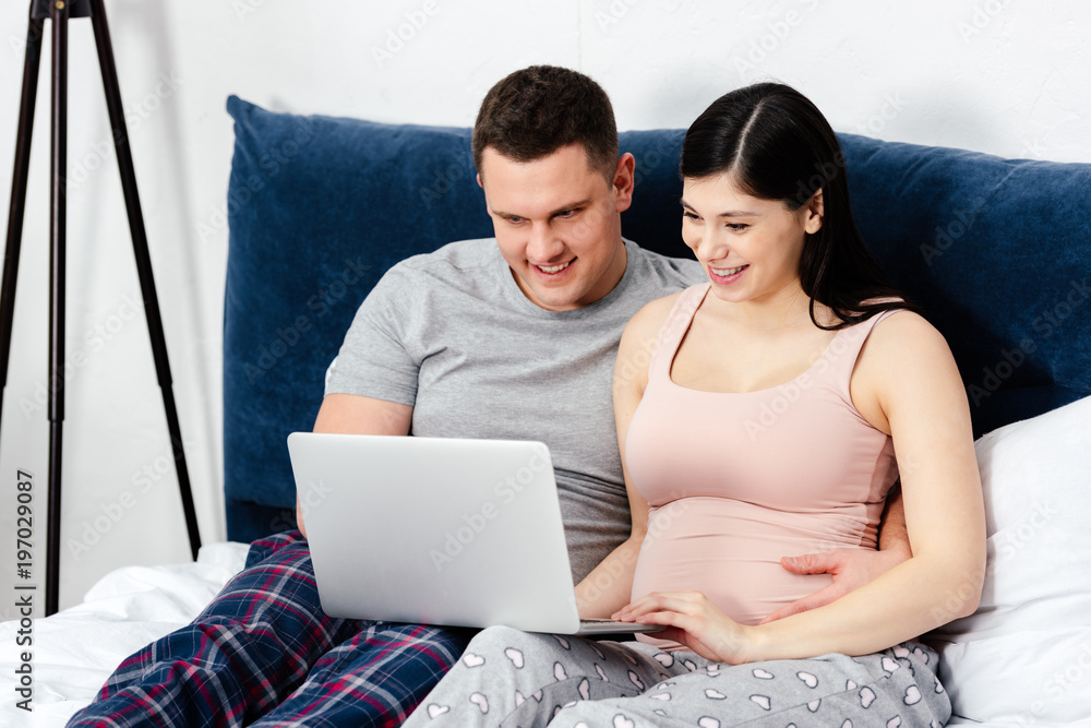 happy young pregnant couple using laptop together on bed