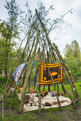 skins arranged below a tent of branches arranged in a wigwam shape viewed low angle outdoors in a clearing in a forest