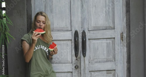 blond handsome girl or woman eating watermelon near her house, backyard, loving frints, having break for snack, vacation in tropical country, holiday mood photo