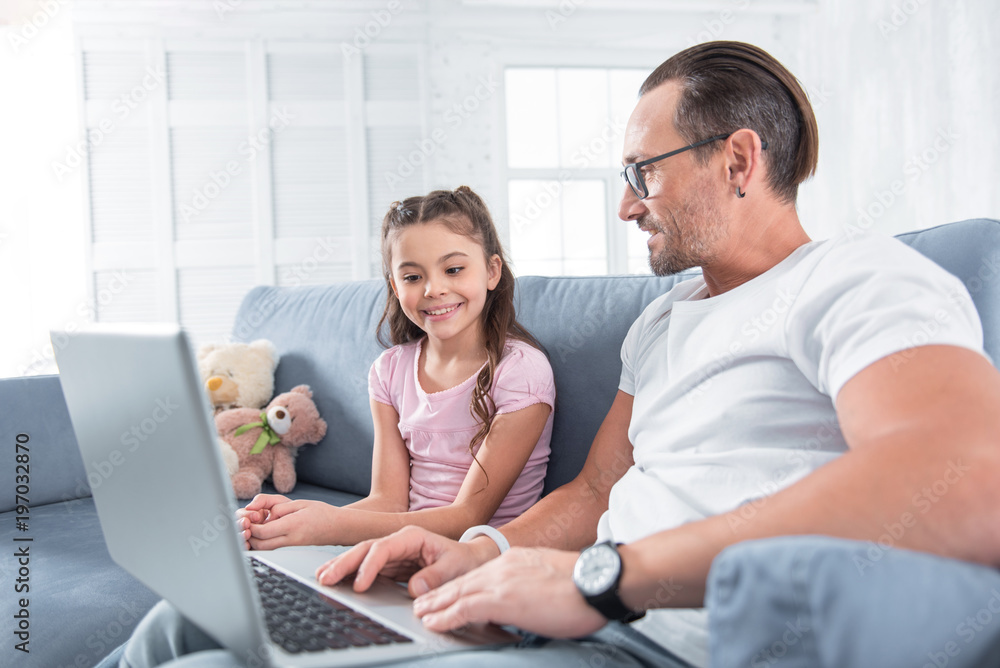 Look here. Happy delighted smart man holding a laptop and showing it to his daughter while being in a great mood Look here. Happy delighted smart man holding a laptop and showing it to his daughter while being in a great mood