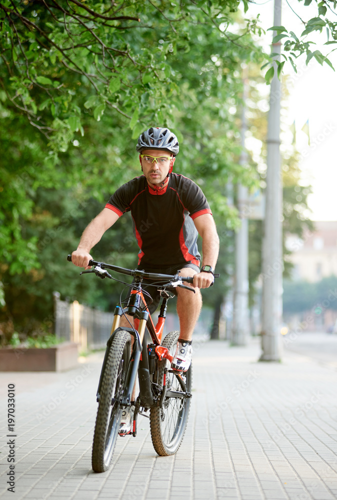 Young bicyclist in cycling sportswear riding on bike near green park. Cyclist shooting for clothing advertisement campaign. Concept of healthy lifestyle, sport advertising, outdoor activities