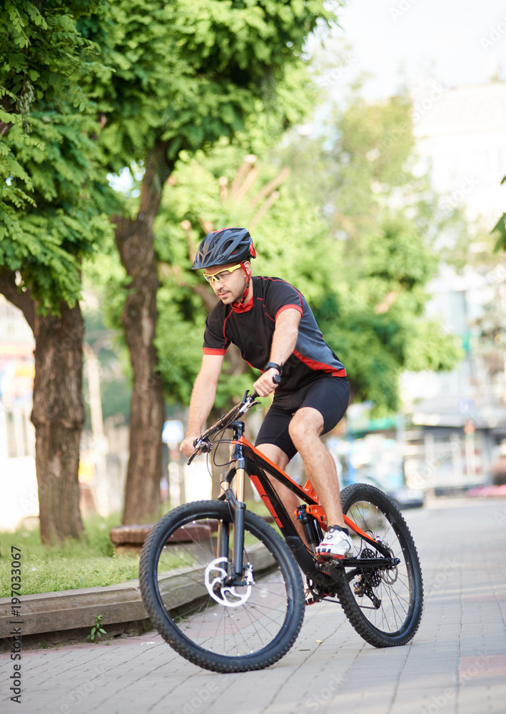 Young male cycler in cycling sportswear and protective gear performing tricks on city streets, getting ready for contest. Cyclist training, improving skills, reaching goal. Concept of motivation Young male cycler in cycling sportswear and protective gear performing tricks on city streets, getting ready for contest. Cyclist training, improving skills, reaching goal. Concept of motivation