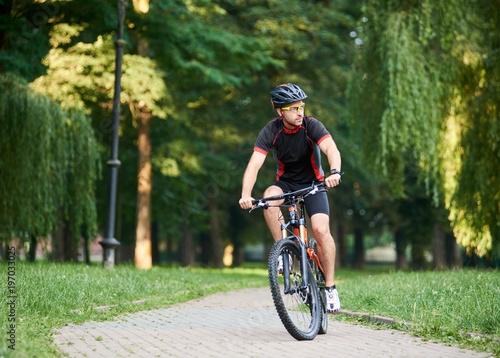 Fototapeta Naklejka Na Ścianę i Meble -  Young man cyclist in professional garment looking to side while riding bicycle down park alley on summer day. Sportsman training thinking about future win in contest. Exercising, reaching goal