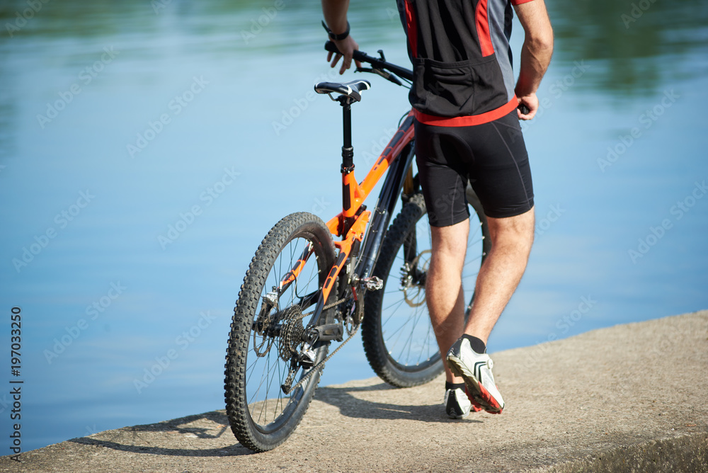 Fototapeta premium Back crop view of guy bicyclist wearing cycling clothes walking next to bicycle down pier to city lake. Sportsman resting and relaxing after training and exercising outdoors. Healthy lifestyle, nature