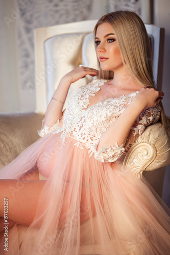 Pretty blonde pregnant young girl with fashion makeup is wearing luxury pink lingerie dress at home in cozy interior with bouquet of roses flowers