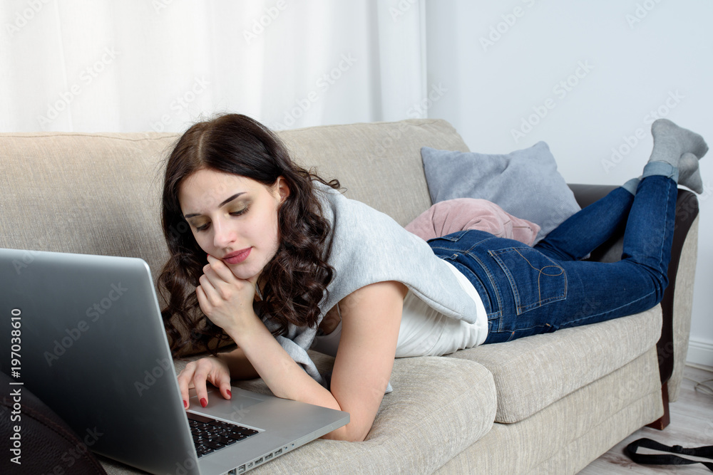 Young woman lying on the couch, works on her laptop computer. Home interior, furniture. Young woman lying on the couch, works on her laptop computer. Home interior, furniture.