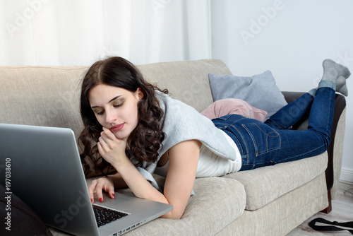 Young woman lying on the couch, works on her laptop computer. Home interior, furniture.
