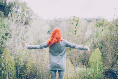 Redhead girl wearing a grey coat standing on the edge of the hill in front of the forest of the mountain and raising her hands. Concept (idea) of freedom, meditation, and motivation.