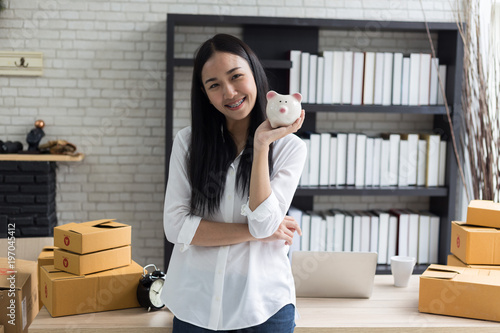 Portrait of smiling asian young woman with piggy bank and cardboard boxes standing in house office,startup Online Marketing concept