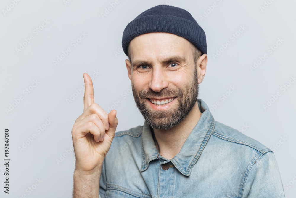 Naklejka premium Attractive middle aged male raises fore finger as gets good idea in mind, wears fashionable clothing, has pleasant smile. Businessman keeps finger up, happy to remember new successful investing plan